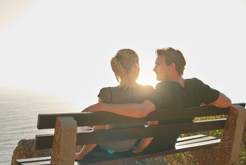 Shot of a loving couple sitting on a bench overlooking the ocean