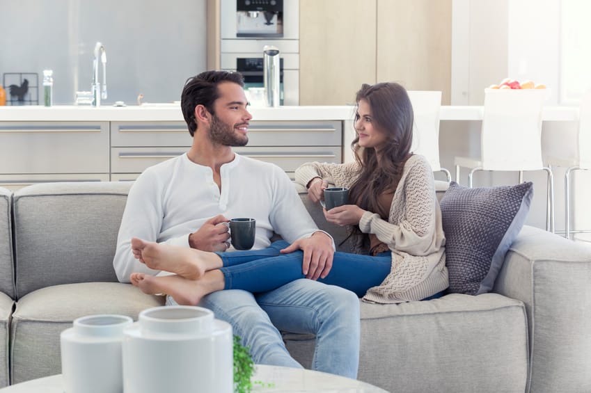 Couple drinking coffee on the sofa. They are talking. They are attractive. There is a modern kitchen I the background.