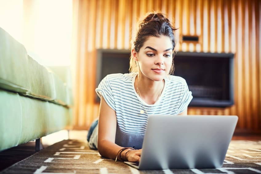 Concentrated woman using laptop at home while lying on the floor. Female blogger is resting on carpet. Hipster is wearing casual.