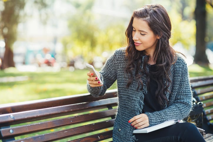 Pretty young girl texting on a smart phone while sitting on a bench in the park.