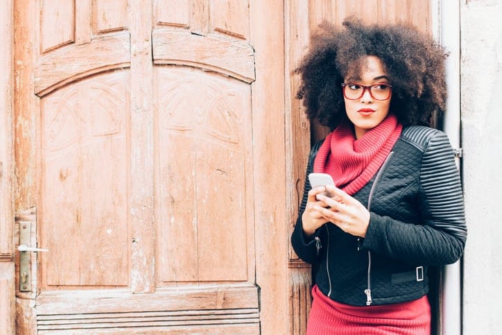 Pensive young woman holding smart phone on street. Front view of head and shoulders. Woman with curly hair, red eyeglasses, red pullover and black jacket. Standing in front of a wooden door.