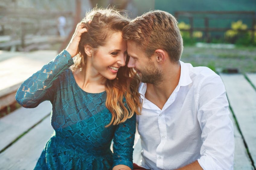Nice couple sitting on a wooden path, outdoor. Girl putting her hand on her hair. Couple looking at each other and smiling, very close. Woman wearing blue dress and man wearing white shirt and claret trousers. Waist up, closeup. Sunshine