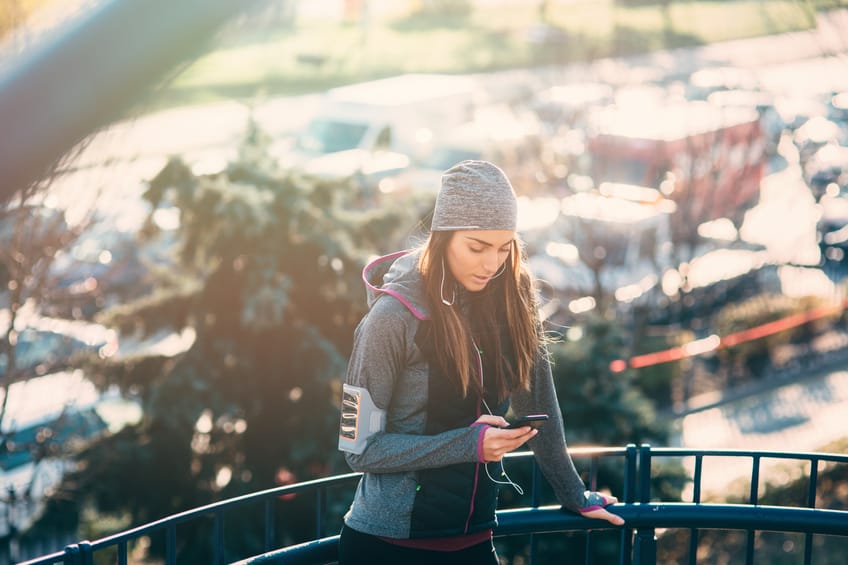 Young beautiful woman resting after the jogging in the city. She is texting on the phone and listening to the music.