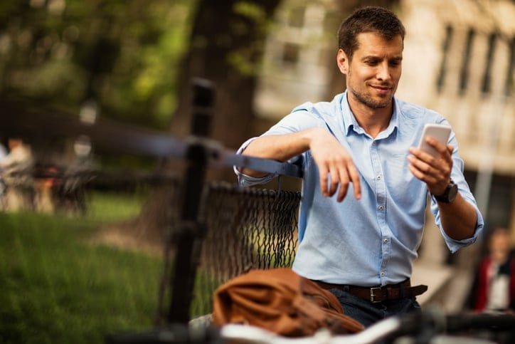 Picture of a young business man who stopped on his bike to look at something peculiar on his smartphone. He is wearing a blue shirt with the sleeves rolled and he is leaning with his arm on a black fence. In the background are a green park and city buildings.