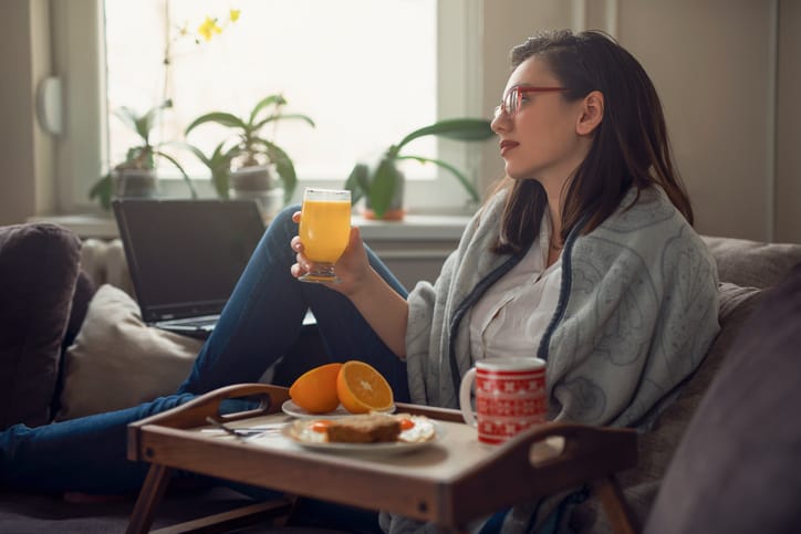 Beautiful young woman sitting casually on the couch, drinking orange juice; a tray with breakfast next to her