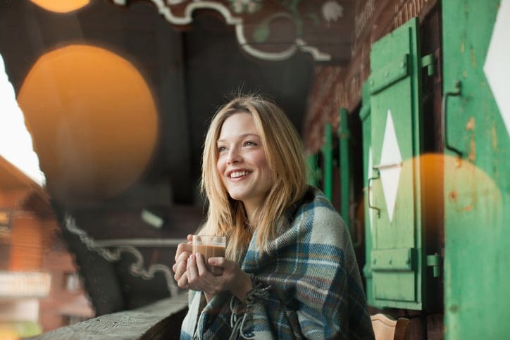 Smiling woman wrapped in a blanket and drinking coffee on cabin porch