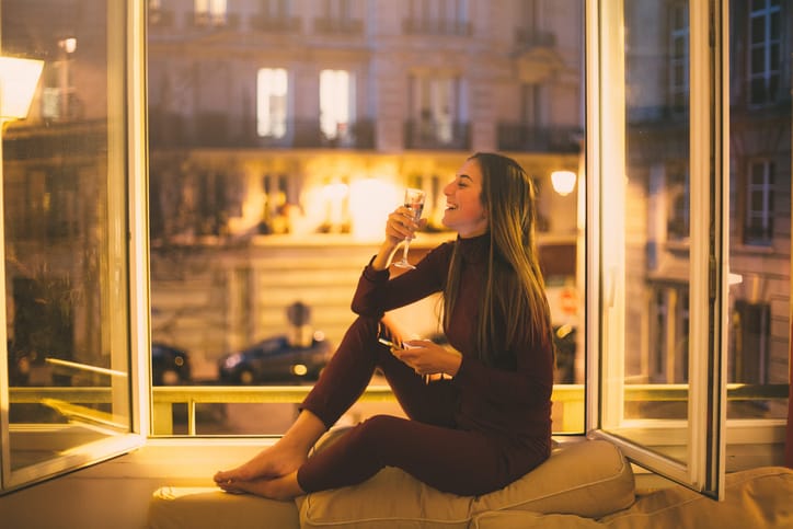 A young woman texting on the phone and drinking wine at home.