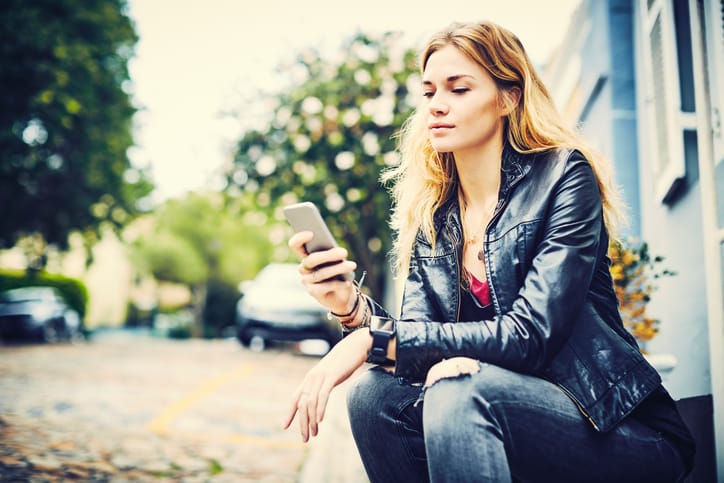 Beautiful woman using smart phone. Young female is text messaging through mobile phone. Hipster is wearing casuals while sitting on roadside.