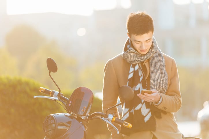 young asian man with mobile phone on motorcycle on road in modern city