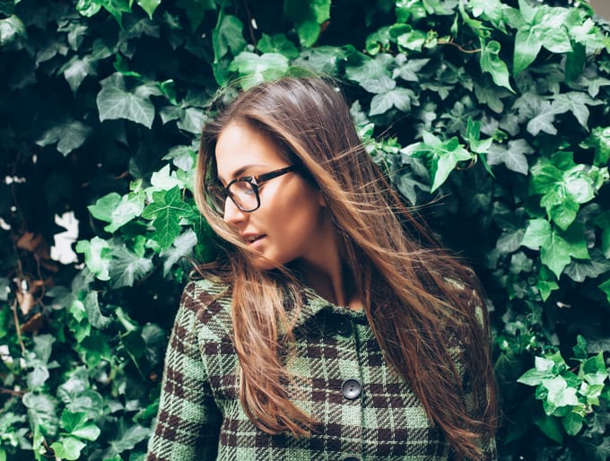 Beautiful young woman standing in front of an ivy wall. Wears eyeglasses and checkered coat. Looking away.
