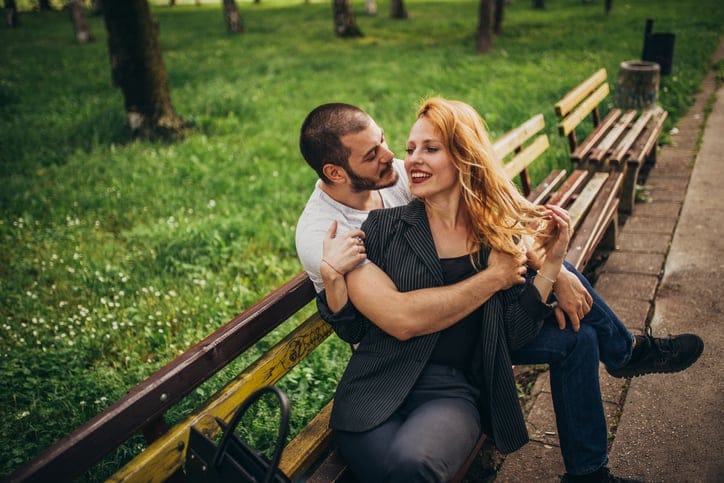 Young Woman and her boyfriend sitting on bench in the park on a sunny day. They man is hugging the woman.