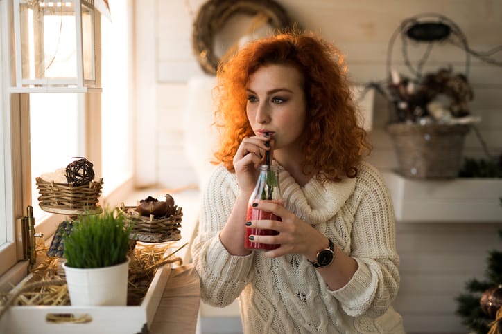 Pensive red-haired woman drinking fruit smoothie while looking away.