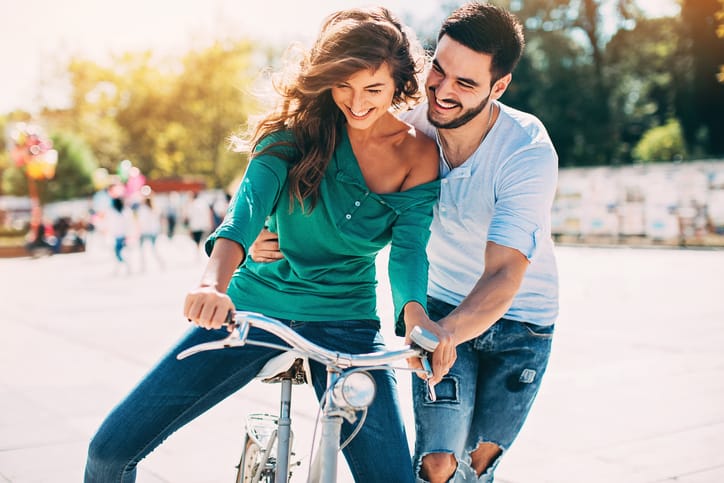 Young man helping his girlfriend riding a bike, outdoors at sunny location.