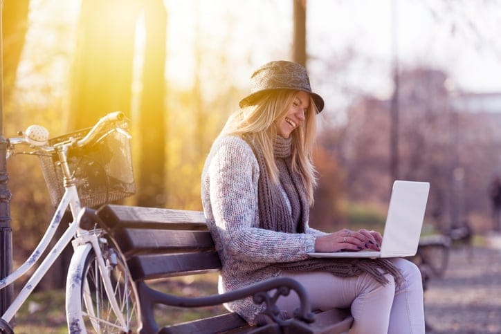 Beautiful blonde girl with hat sitting on the bench working on laptom. Wi fi in the citty.