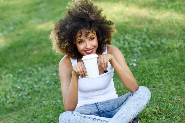 Beautiful young African American woman with afro hairstyle. Girl drinking coffee with a take away glass in park sitting on grass wearing casual clothes smiling.