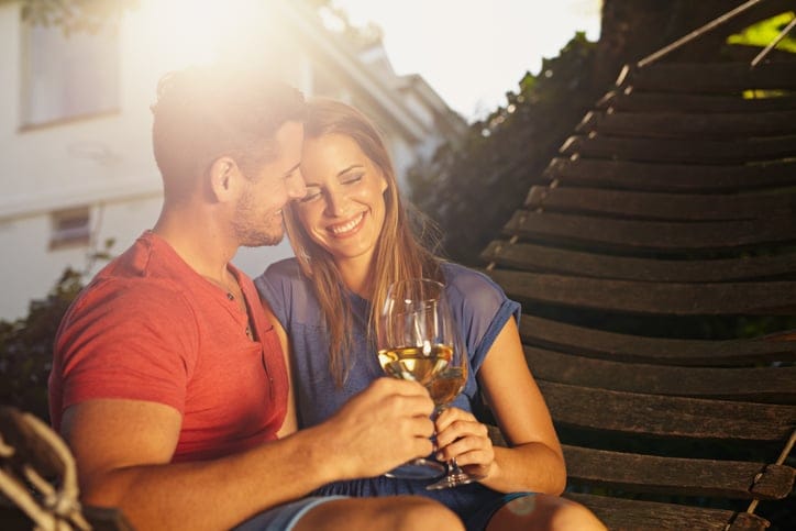 Beautiful young couple toasting wine outdoors. They are sitting on a hammock smiling and drinking wine with bright sunlight in backyard. Young man and woman spending quality time together on weekend.