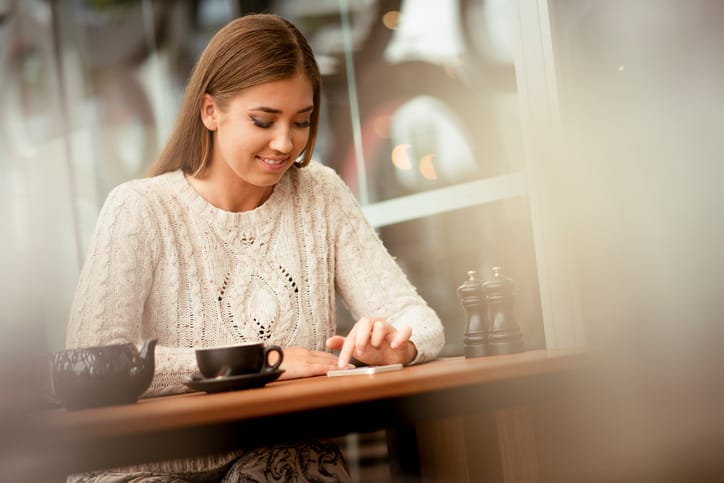 Young woman sitting in a cafe in the city. She is using her mobile phone, looking at it and she is smiling. There is also a cup of tea on the table.