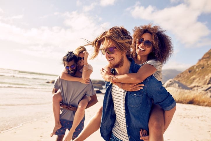 Two happy young men giving their girlfriends piggyback rides. Group of young people enjoying themselves during summertime at the beach.