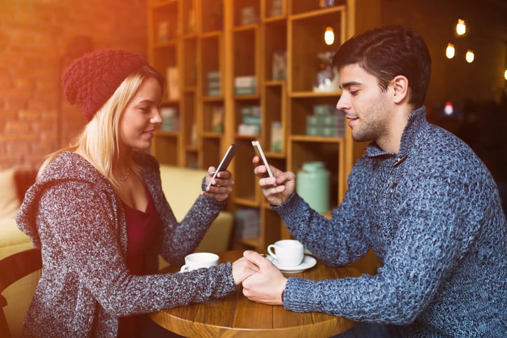 Young couple is sitting in a coffee shop holding hands and text messaging on the mobile phones at the same time without