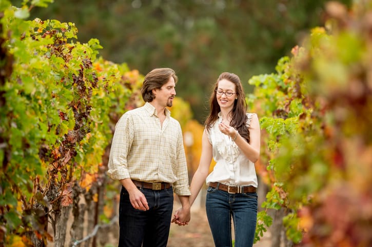 A heterosexual couple holding hands and walking together on a path looking at the grapes at a vineyard in the Napa Valley while on their honeymoon vacation.