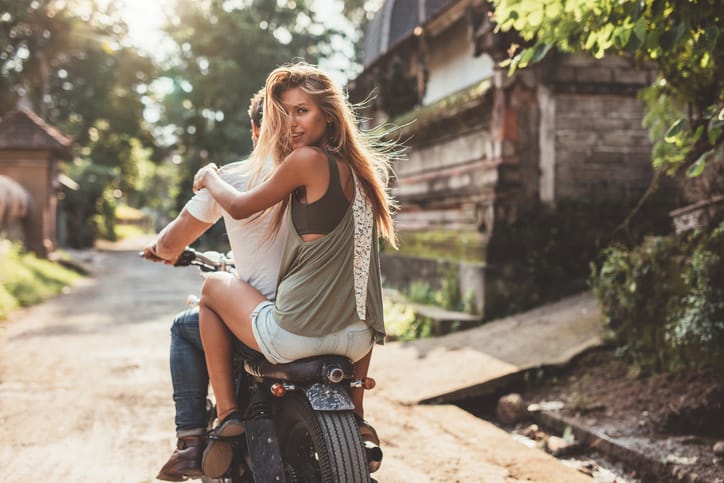 Rear view shot of attractive young woman sitting on back of her boyfriend riding bike through village road.