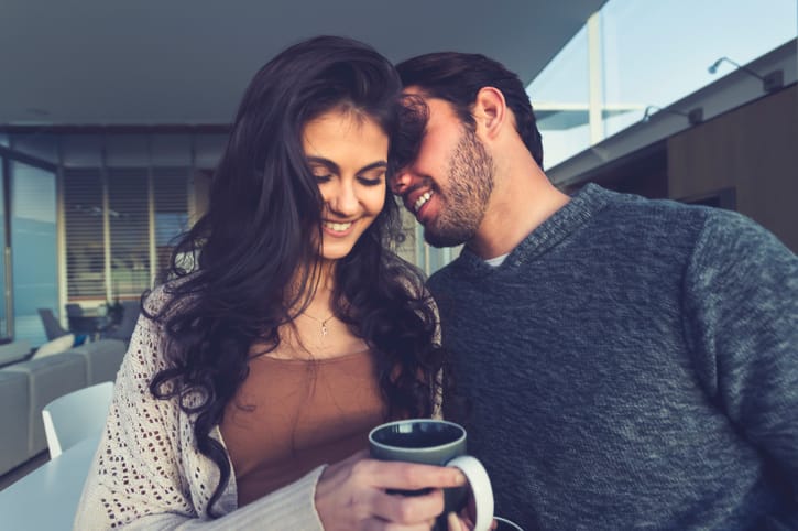 Couple flirting playfully. He is snuggling into and is about to kiss her on the neck. He may be whispering a secret. She is smiling and happy. Close up.