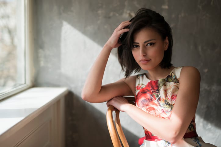 Fashion portrait of a beautiful black-haired woman sitting on a chair by the window.