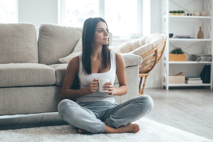 Attractive young woman sitting on the carpet at home and holding coffee cup