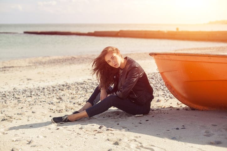 A girl sideways sitting near a red boat on the beach by the sea with beautiful sunset on the background. Woman in jeans, pullover and jacket on the sand