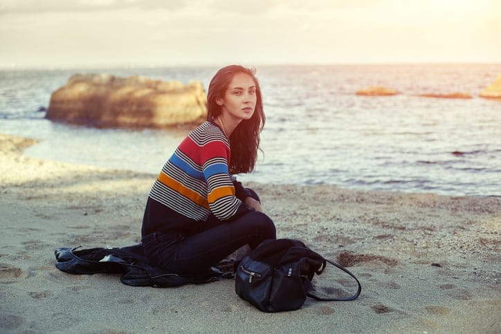 A girl sitting on the sand beach by the sea with beautiful sunset on the background. Woman in jeans, pullover and jacket on the sand