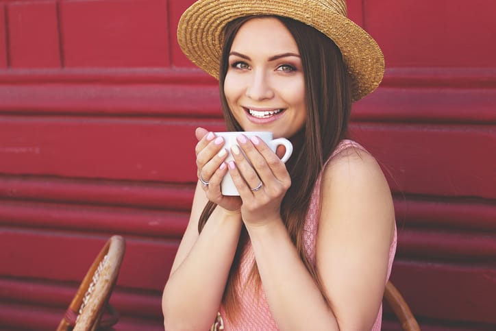 Happy coffee time. Closeup portrait of beautiful young smiling woman in pink dress and hat keeping cup of coffee in her hands while sitting in French vintage cafe.