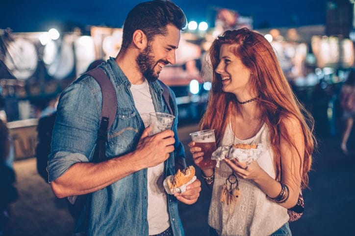 A young couple having snack and drink at an outdoors music festival. Drinking beer and eat burgers. Night with sparkling lights in back ground.