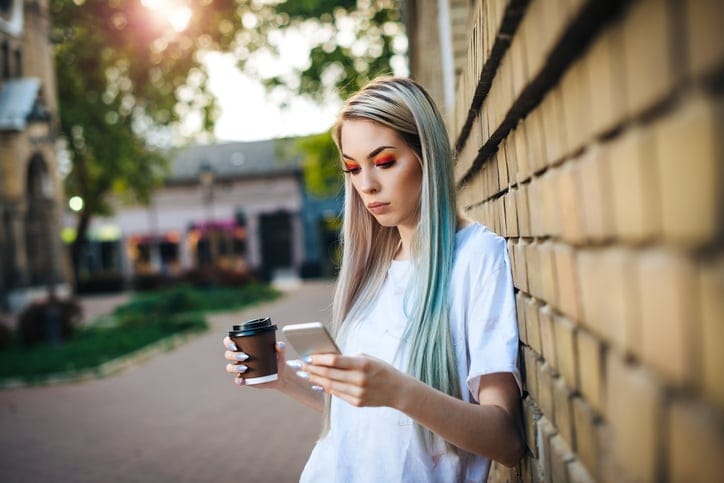 20 year old hipster woman dressed in street style clothing living typical London urban lifestyle. Woman is leaning on old brick wall and using phone for communication and music.