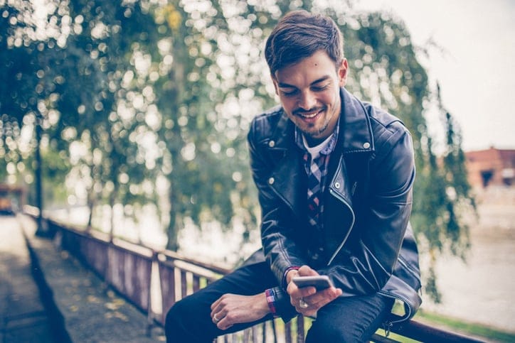 Young man using phone while sitting on a fence in city park