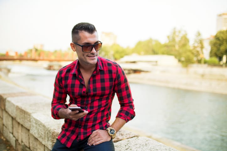Handsome young guy sitting by the river and using mobile phone