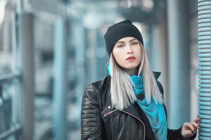cool woman in leather jacket and black cap posing in the city, wearing blue scarf and looking at camera.