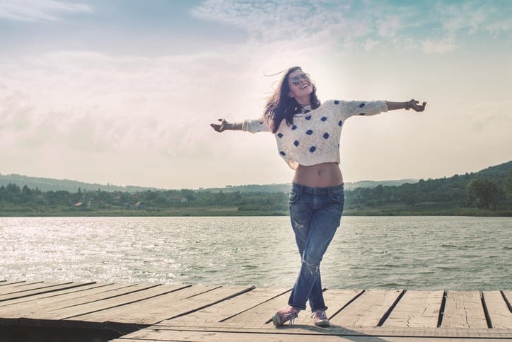 Young woman with open arms, standing on a dock by the lake, smiling
