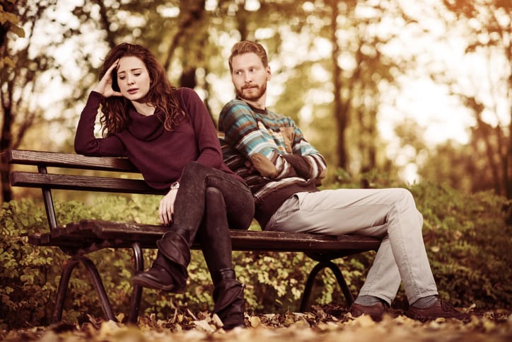 Young displeased couple with problems in their relationship sitting on a bench in autumn park. Focus is on woman.