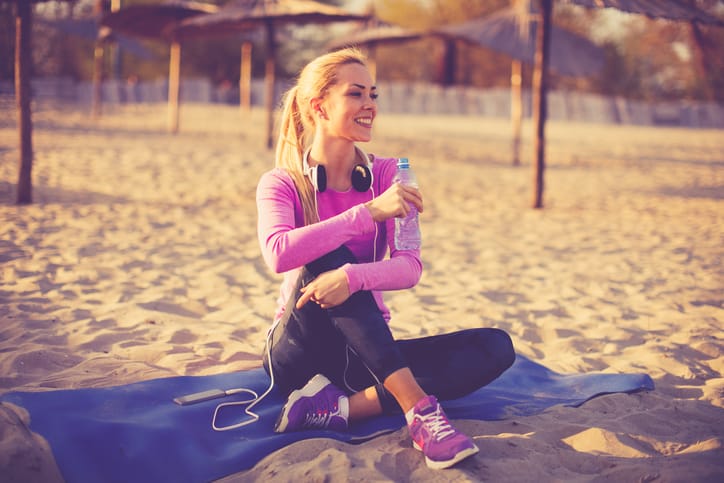 Beautiful woman resting after jogging on the beach at sunset and holding bottle of water