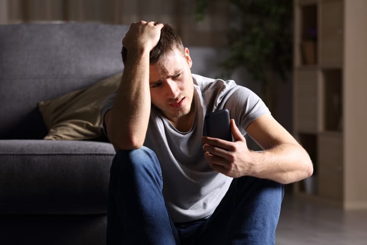 Single sad man checking mobile phone sitting on the floor.
