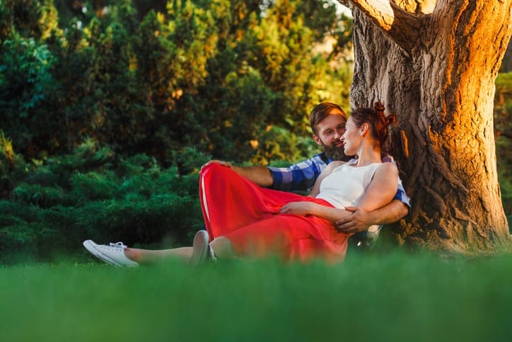 A beautiful couple is resting outside the city in park, sitting near a big tree in the garden look at each other and smiling. Love story
