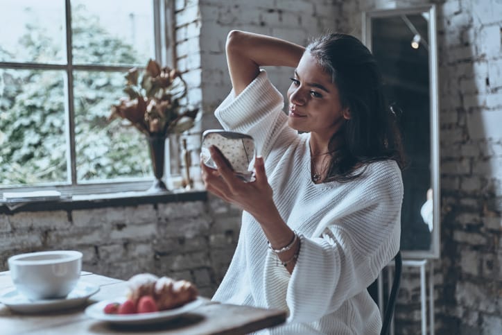 Young woman sitting at breakfast table, looking at herself in pocket mirror