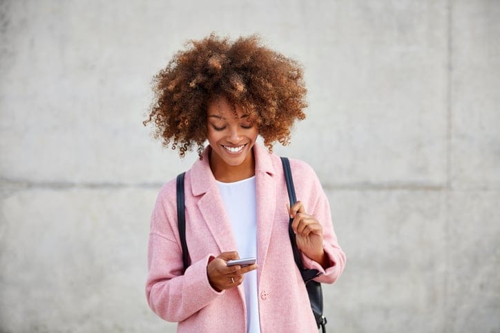 Happy young woman text messaging through smart phone. Beautiful female is with frizzy short hair. She is wearing pink overcoat.