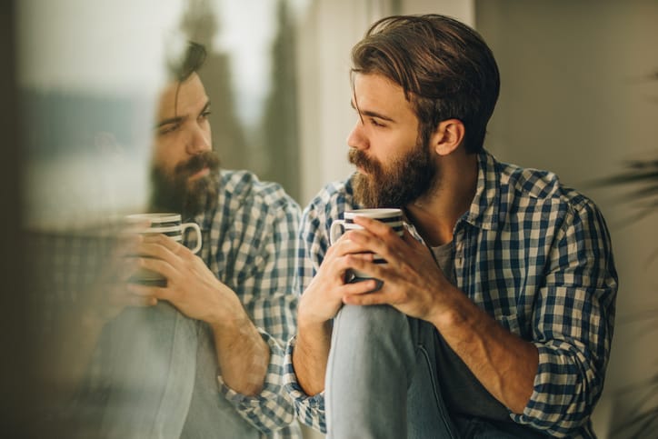 Young man sitting by the window during his coffee time and looking through it. His reflection is seen in window.
