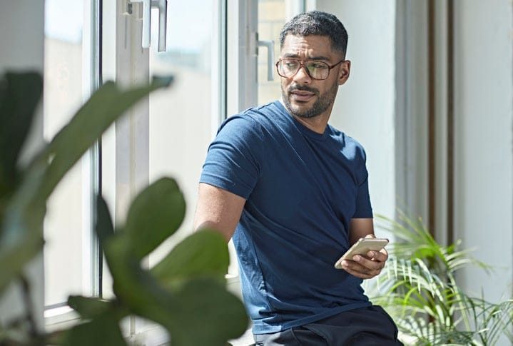 Mixed race male weekend worker looking out the window with concerned expression after learning bad news on smart phone.