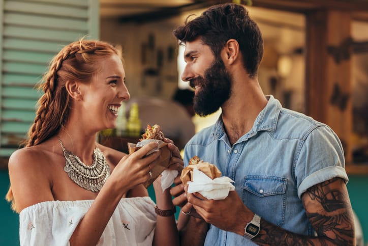 Couple eating sandwiches smiling at each other.