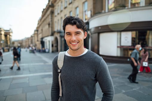 Young man looking at the camera with a big smile while in the city street. He is carrying a shoulder bag.