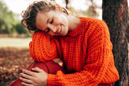 Outdoor image of a attractive young woman smiling, wearing orange knitted sweater posing on fall nature background. Beautiful female with closed eyes has dreamy expression, resting outdoor in the park