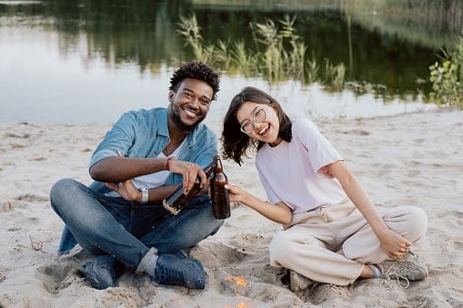 Couple of friends are sitting next to each other on beach by lake smiling laughing woman of Korean Asian beauty is holding bottle of beer in hand is leaning towards man tapping each other cheers