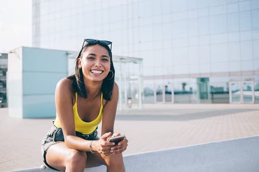 Portrait of toothy rejoice hipster girl in stylish clothing holding modern cellphone gadget in hands and resting on urban setting with public internet, prosperous female blogger with mobile phone